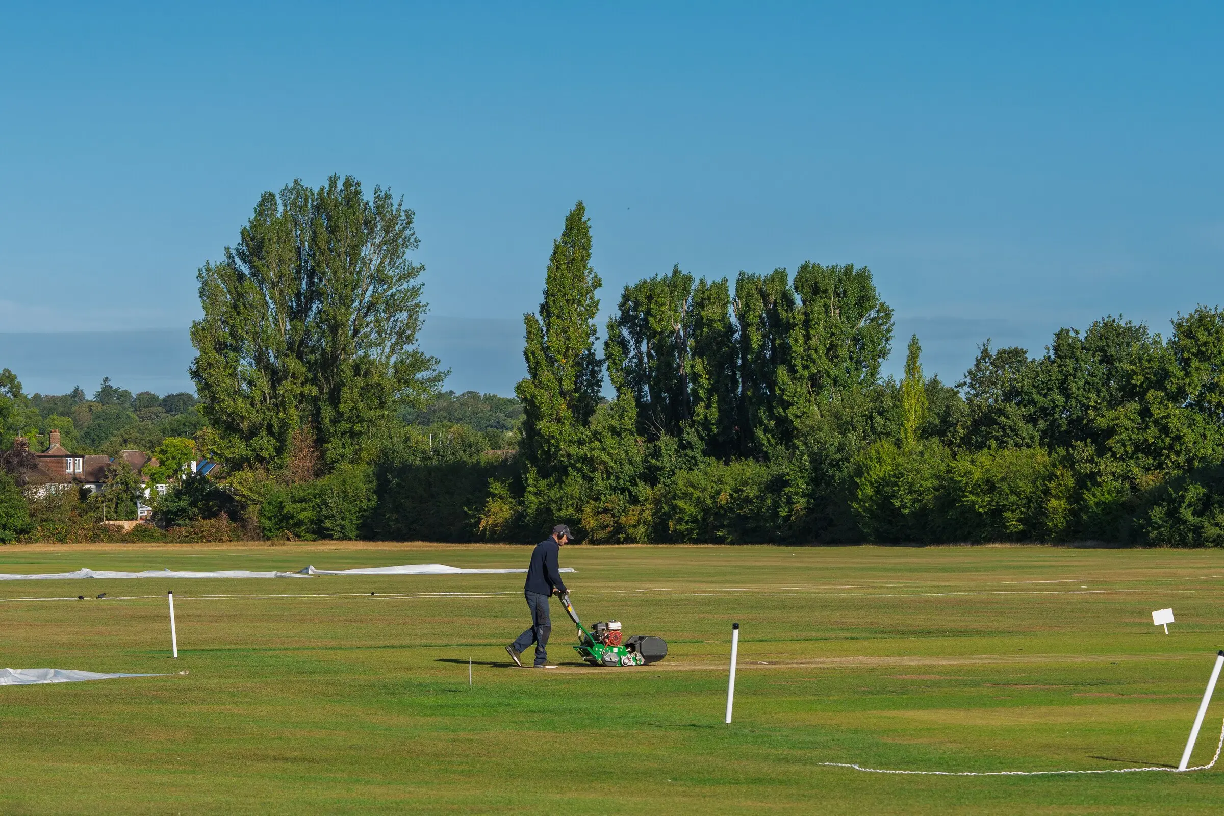 A groundsman mowing a large sports outfield with boundary markers and pitch covers laid out